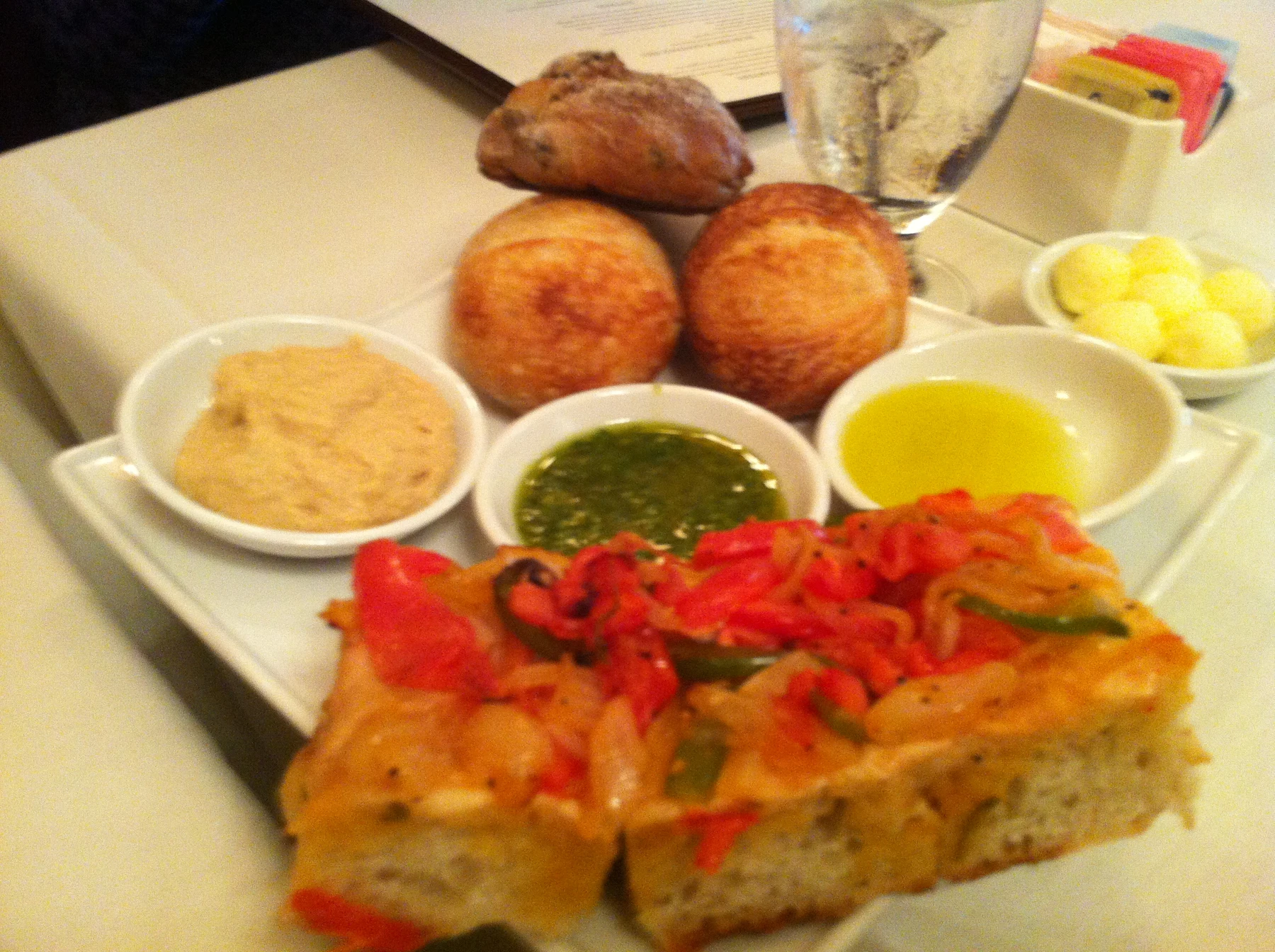 Focaccia topped with red vegetables beside rolls and dipping sauces