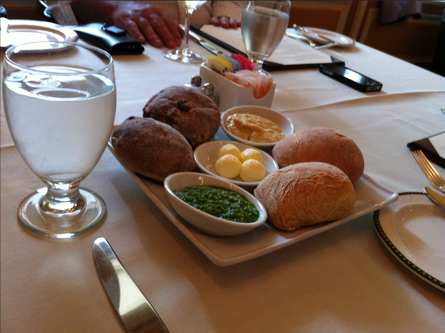 Textured crusty rolls, vivid green pesto, butter and hummus beside a water goblet at 1792