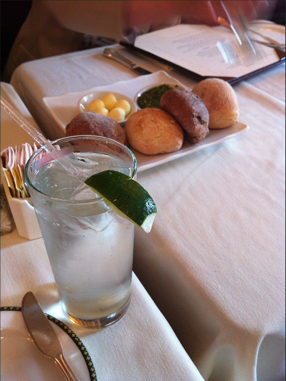 A glass with ice and a lime wedge in sharp foreground focus, with bread rolls, butter and pesto on the table behind